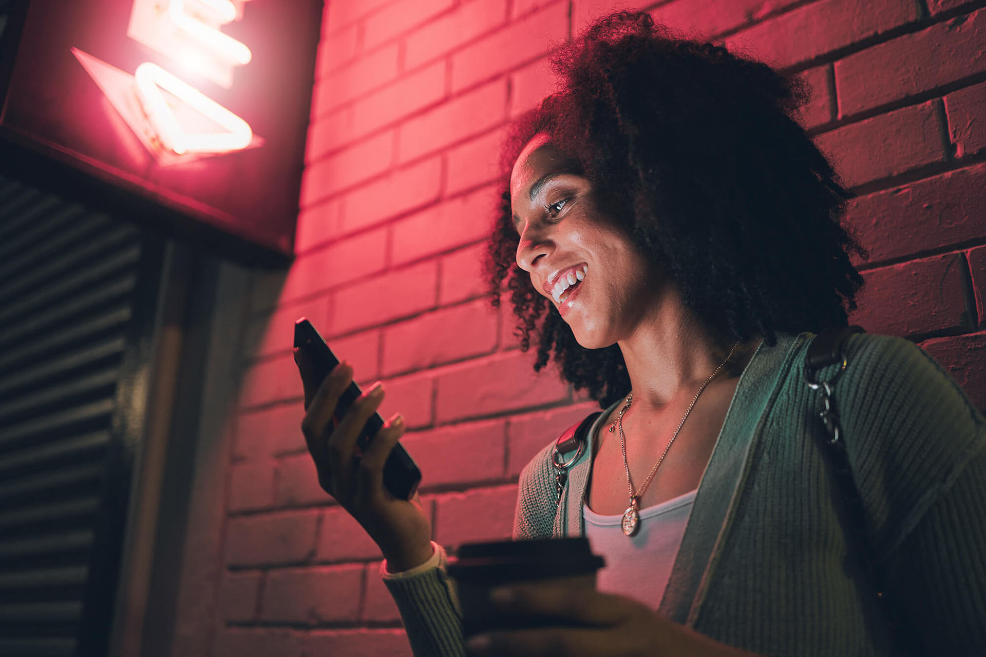 Afro-american woman checking social media management on mobile phone by a brick wall, under the red neon light.