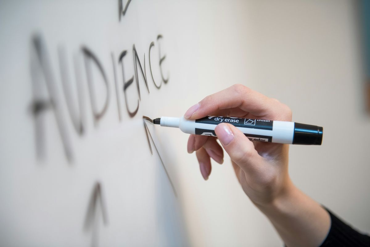 Person writing "AUDIENCE" on a whiteboard with a marker. An important part and key differentiation in digital product design development.